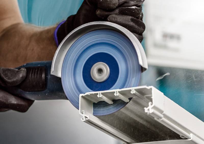 Eine Person benutzt ein Elektrowerkzeug mit einer rotierenden Kreissäge, um ein weißes Metall- oder Kunststoffobjekt zu schneiden, wobei sie Schutzhandschuhe trägt.