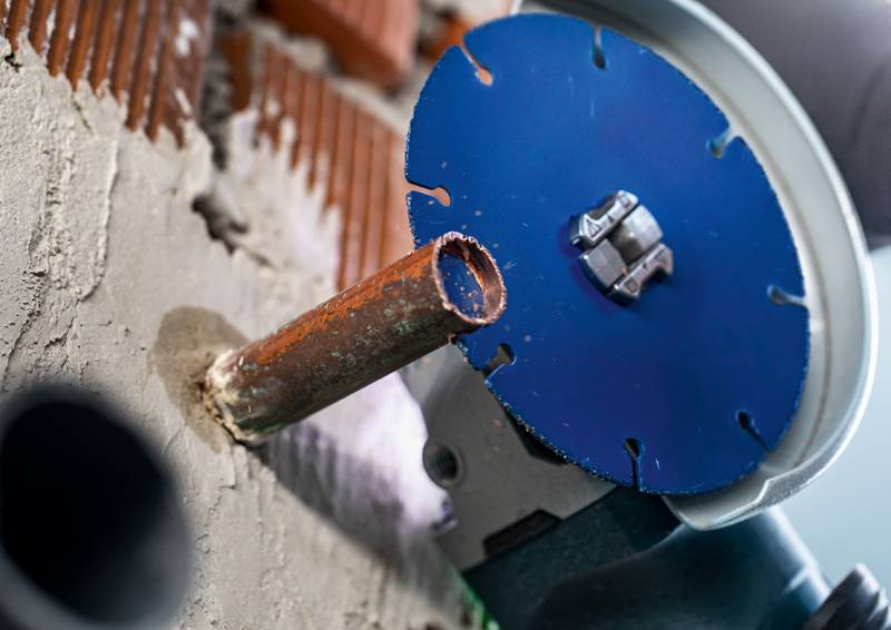 Eine Nahaufnahme einer blauen Kreissäge, die ein Metallrohr durchtrennt, das aus einer Betonwand hervorsteht, mit sichtbaren Fliesen im Hintergrund.