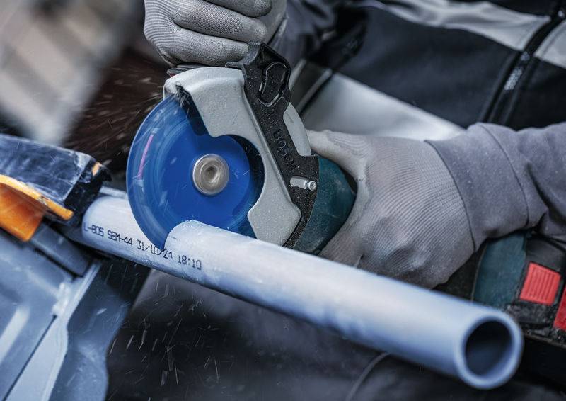 Eine Person mit Handschuhen benutzt eine Elektrosäge, um ein Kunststoffrohr zu schneiden, wobei Staubpartikel in der Luft um den Schnitt herum sichtbar sind.