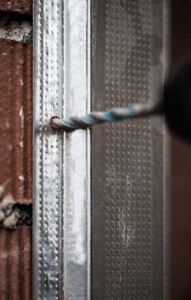 „Eine Nahaufnahme eines Bohrers, der ein Loch in einen Metallrahmen bohrt, der an einer Ziegelmauer befestigt ist, wobei Konstruktions- oder Installationsarbeiten hervorgehoben werden.