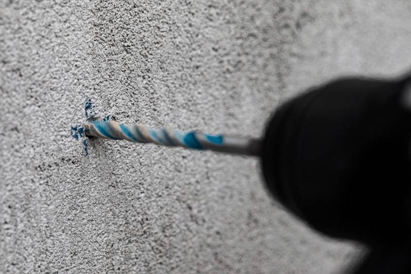 Eine Nahaufnahme eines Bohrers mit einem blau-silbernen Aufsatz, der in eine graue Betonwand eindringt und eine Heimwerker- oder Bauaufgabe zeigt.