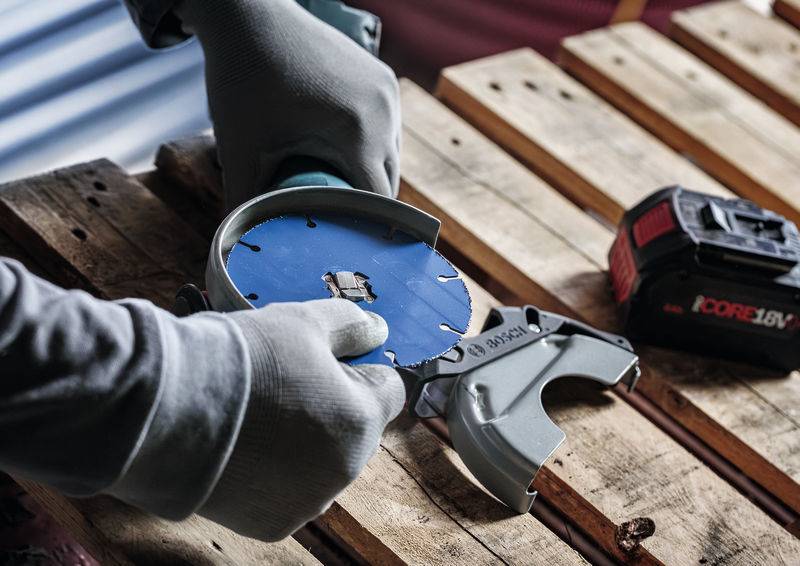 Hände mit Handschuhen halten einen kreisförmigen Bohraufsatz auf einer Holzoberfläche, neben einer Bosch-18V-Batterie, was auf eine Holzbearbeitungsaufgabe hindeutet.