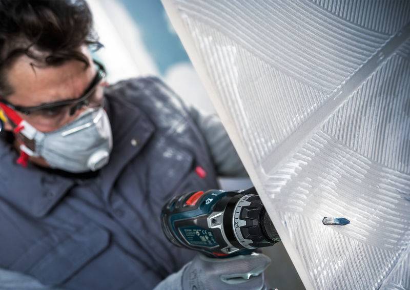 Eine Person benutzt eine Bohrmaschine auf einer weißen Oberfläche und trägt dabei eine Schutzbrille, eine Maske und Handschuhe, was den Fokus auf Sicherheit und Präzision bei der Arbeit unterstreicht.