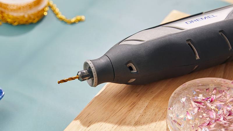 Ein Elektroschleiffer mit einem Bohraufsatz liegt auf einer Holzoberfläche, umgeben von Bastelmaterialien, was auf eine kreative Werkstatt hindeutet.