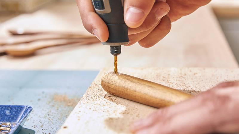 Eine Hand benutzt einen Elektroschrauber an einem Holzdübel, wobei Sägespäne auf einer Werkbank entstehen, was auf einen Holzbearbeitungsprozess hinweist.