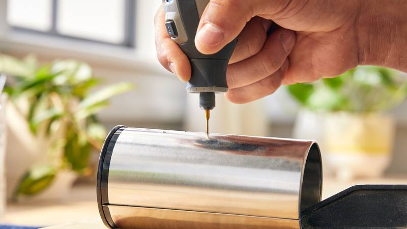 Eine Person nutzt ein Drehwerkzeug, um ein Design auf eine glänzende zylindrische Metalloberfläche zu gravieren, mit verschwommenen Pflanzen im Hintergrund.