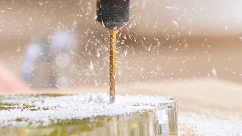 Ein Bohrer schneidet in eine Glasoberfläche ein und erzeugt feines weißes Pulver und Partikel, die in alle Richtungen davon spritzen.