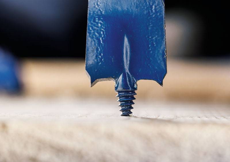 Eine Nahaufnahme eines blauen Bohrers mit flacher Spatenspitze und Schraubenpunkt, der in eine Holzoberfläche eindringt und Textur sowie Detail zeigt.