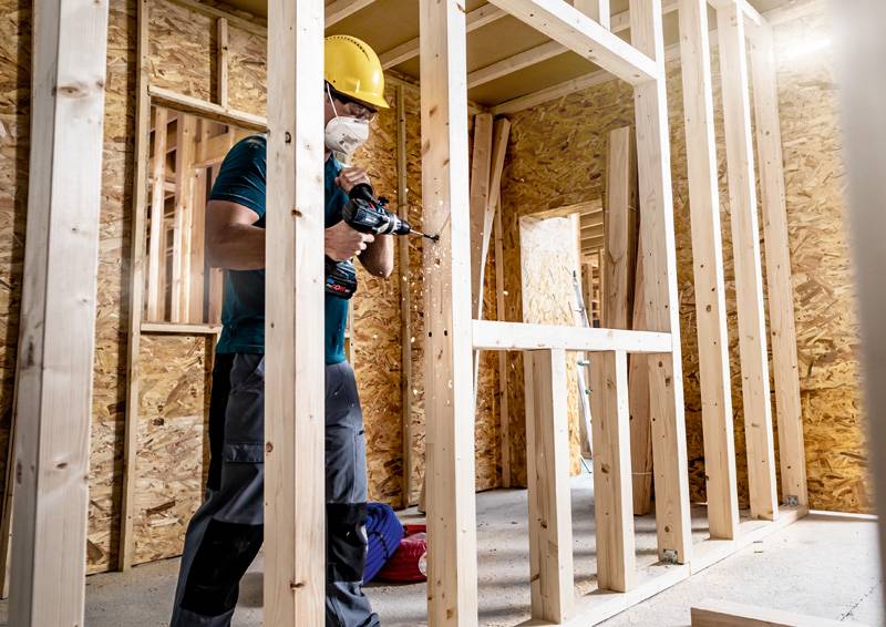 Ein Bauarbeiter mit Helm und Maske benutzt eine Elektrobohrmaschine, um einen Holzrahmen in einem sich im Bau befindlichen Gebäude zusammenzubauen.