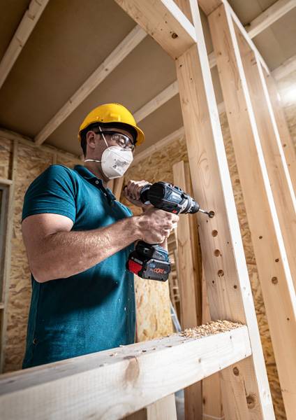 Ein Bauarbeiter mit Schutzhelm und Gesichtsmaske verwendet einen Elektroschrauber an einem Holzrahmen auf einer Baustelle.