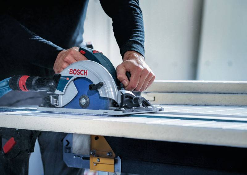 Eine Person im schwarzen Hemd bedient eine Bosch-Kreissäge und schneidet eine weiße Platte auf einer Werkbank in einer Werkstatt durch.