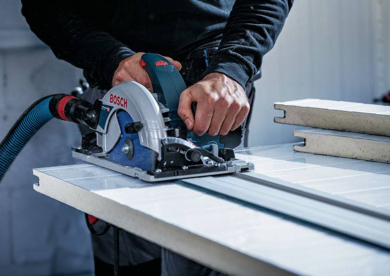 Eine Person verwendet eine Bosch-Kreissäge zum Schneiden einer weißen Platte in Innenräumen, wobei zwei gestapelte Platten in der Nähe liegen. Die Säge ist an einen Staubabscheider angeschlossen.