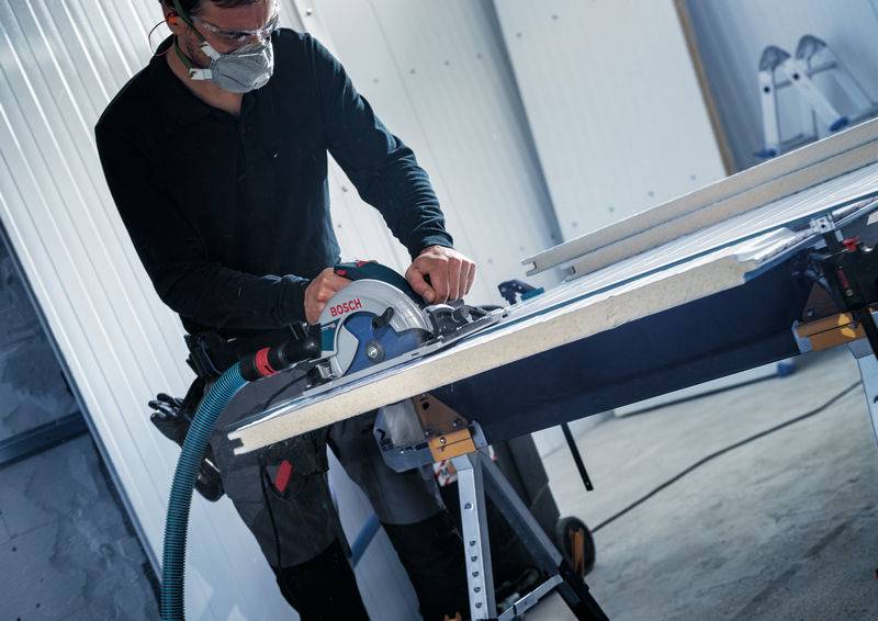 Eine Person in Schutzausrüstung verwendet eine Kreissäge, um ein Materialblatt auf einer Werkbank in einer Werkstattumgebung zu schneiden.
