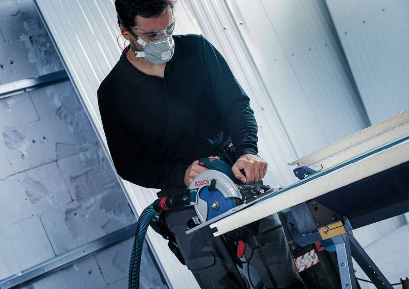 Eine Person, die eine Schutzbrille und eine Maske trägt, benutzt ein Bosch-Elektrowerkzeug zum Zuschneiden von Holz auf einer Werkbank in einer Werkstatt.