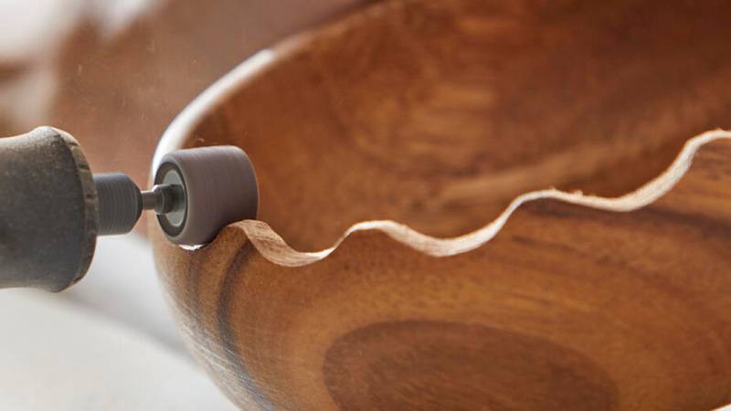 Eine Person benutzt ein Elektroschleifergerät, um die gewellte Kante einer hölzernen Schale abzuschleifen und deren Oberfläche für ein poliertes Finish zu glätten.