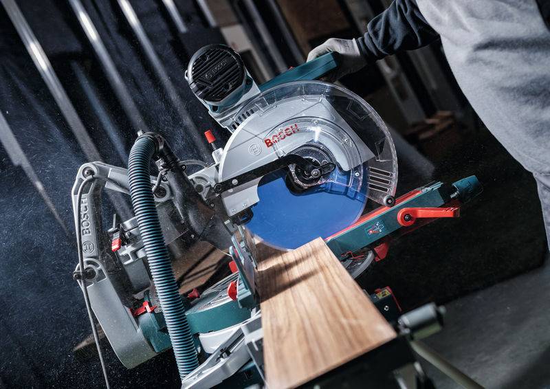 Eine Person bedient eine Bosch-Gehrungssäge und schneidet ein Holzbrett. Die Säge ist in einer Werkstattumgebung positioniert, wobei Sicherheitsvorkehrungen beachtet werden.