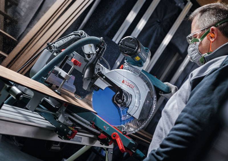 Eine Person bedient eine Elektrosäge und trägt Schutzausrüstung einschließlich Schutzbrille und Maske in einer Werkstattsituation.