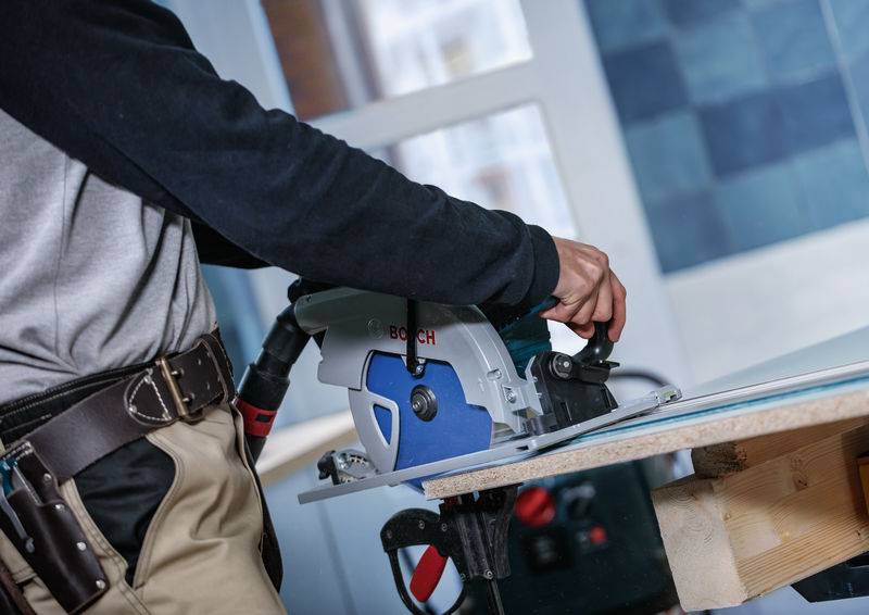 Person, die eine Bosch-Kreissäge verwendet, um ein weißes Brett auf einer Werkbank in Innenräumen zu schneiden, trägt einen Werkzeuggürtel und ein schwarzgraues Hemd.