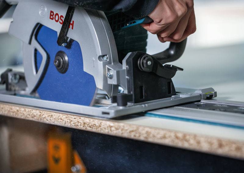Eine Person, die eine Bosch-Kreissäge verwendet, um ein Holzstück zu schneiden, mit Fokus auf das Sägeblatt und das Holz, was eine Holzbearbeitungstätigkeit andeutet.