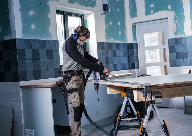 Eine Person arbeitet mit einem Elektrowerkzeug auf einer Arbeitsplatte in einem teilweise renovierten Zimmer und trägt Schutzausrüstung wie Schutzbrille und Gehörschutz.