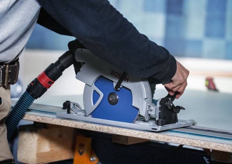 Eine Person nutzt eine Bosch-Kreissäge, um ein Holzstück auf einer Werkbank zu durchtrennen. Die Säge ist mit einem Staubsauger zur Staubabsaugung verbunden.