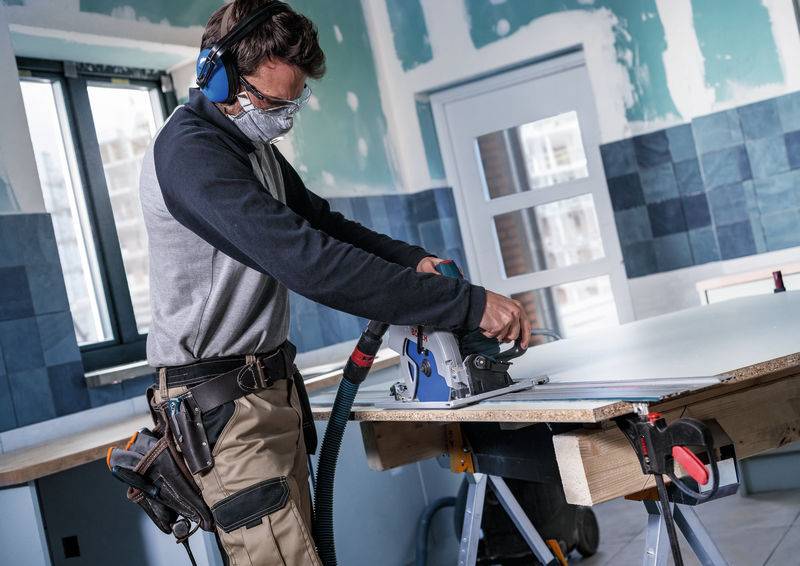 Eine Person in Schutzausrüstung benutzt eine Kreissäge an einem Brett in einem teilweise konstruierten Raum mit gefliesten Wänden und Fenstern.