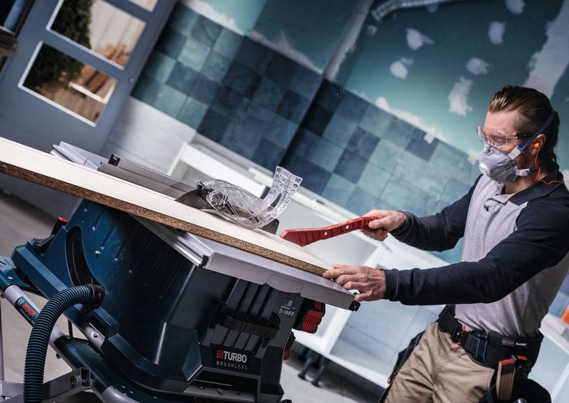 Eine Person in Schutzausrüstung bedient eine Kreissäge auf einer Werkbank in einer Werkstatt und schneidet ein Holzbrett zu.