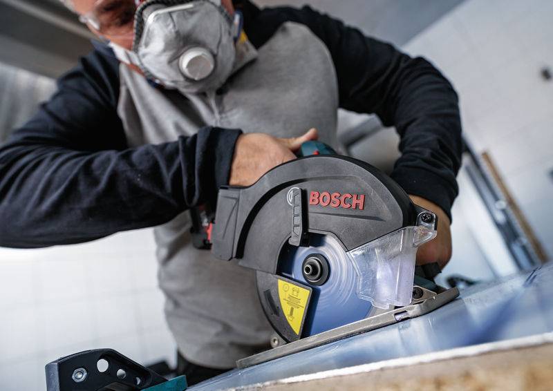 Eine Person, die eine Schutzmaske und eine Schutzbrille trägt, bedient eine Bosch-Elektrosäge und schneidet ein blaues Material in einer Werkstattumgebung.