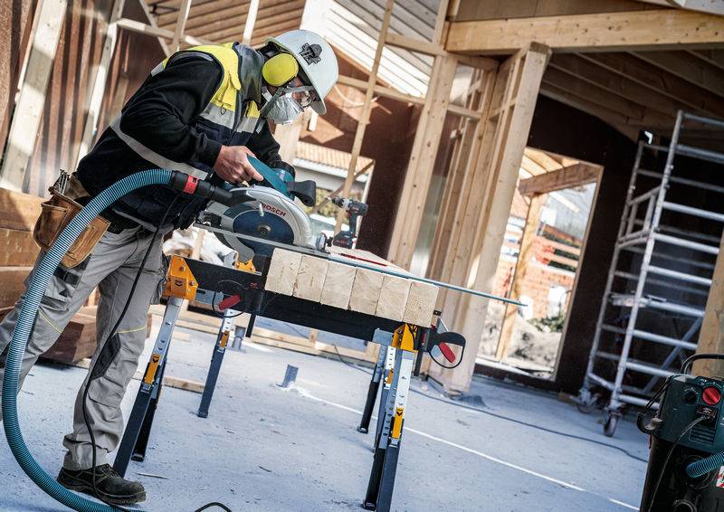 Der Bauarbeiter schneidet Holz auf einem Tisch mit einer Kreissäge zurecht, trägt Schutzausrüstung einschließlich Helm und Maske und arbeitet in einem sich im Bau befindlichen Gebäude.