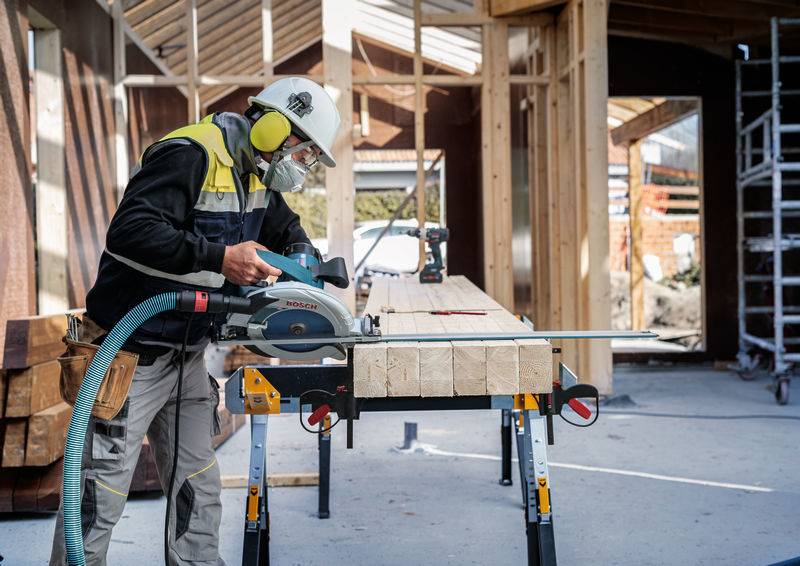 Ein Bauarbeiter in Schutzausrüstung benutzt eine Kreissäge, um Holzbretter auf einer Werkbank auf einer Baustelle zu zuschneiden.