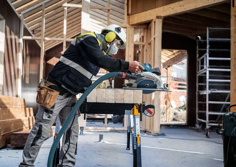 Ein Bauarbeiter mit Schutzausrüstung bedient eine Kreissäge an Holz auf einer Baustelle. Im Hintergrund sind Gerüste und Holzrahmen sichtbar.