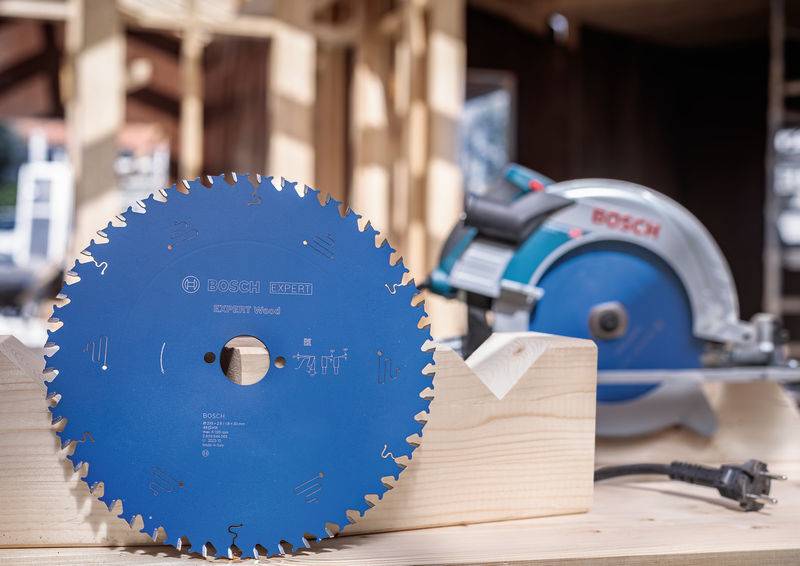 Ein blau gefärbtes kreisrundes Sägeblatt steht aufrecht auf einer hölzernen Tischplatte in einer Werkstatt, mit einer unscharfen elektrischen Säge und Holzbalken im Hintergrund.