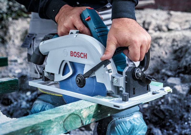 Eine Person, die eine Bosch-Kreissäge hält, schneidet grünes Holz im Freien. Der Fokus liegt auf dem Sägeblatt des Werkzeugs und den Händen, die es führen.