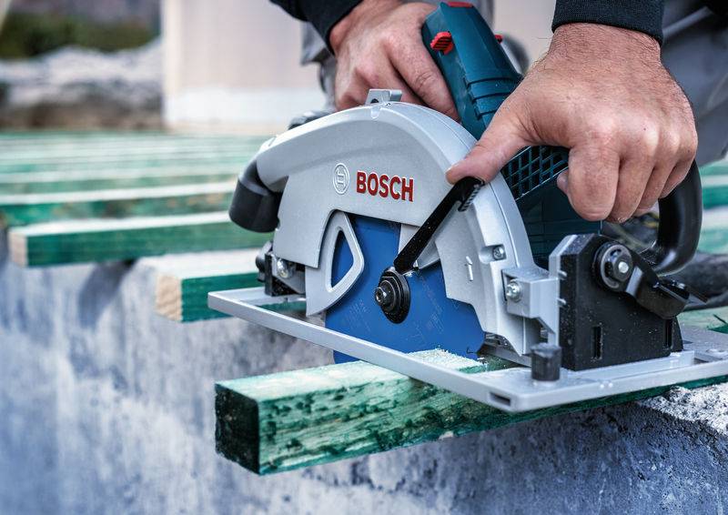 Eine Person benutzt eine Bosch-Kreissäge, um ein Stück grünes Holz auf einer Baustelle zu schneiden. Der Fokus liegt auf Präzision und Holzbearbeitung.