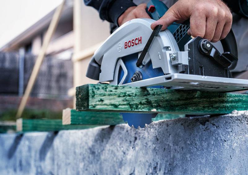 Person schneidet grünes Holzbrett mit Bosch-Kreissäge auf einer Betonoberfläche. Der Arbeiter konzentriert sich auf präzises Schneiden.