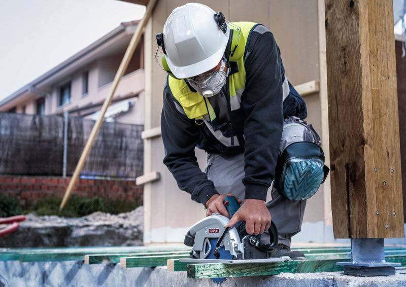 Ein Bauarbeiter, der einen Helm und Schutzausrüstung trägt, benutzt eine Kreissäge, um Holzbretter auf einer Baustelle zu schneiden.
