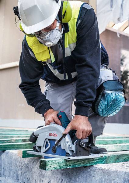 Ein Arbeiter, der Schutzausrüstung trägt, nutzt eine Bosch-Elektrosäge, um ein Stück grünes Holz auf einer Baustelle zu schneiden.