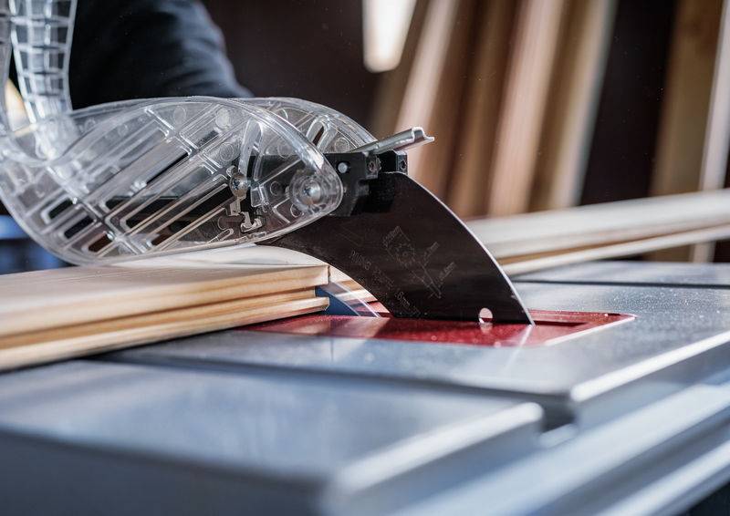 Eine Tischkreissäge schneidet ein Holzbrett mit einer transparenten Schutzvorrichtung, die das scharfe Sägeblatt abdeckt, in einer Werkstattumgebung.
