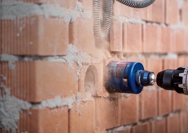 Ein Bohrgerät mit einem blauen Lochsägenaufsatz schafft ein Loch in einer roten Backsteinmauer, umgeben von Staub und Schutt.