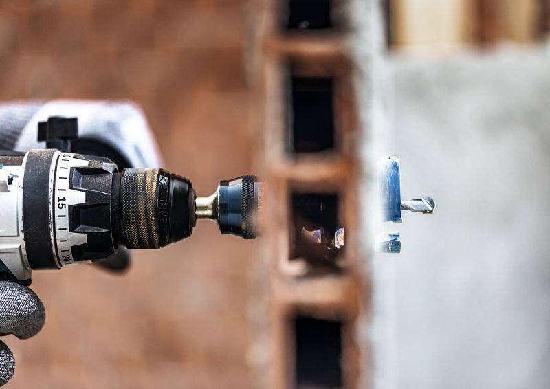 Eine Nahaufnahme einer Elektrobohrmaschine, die in eine Ziegelmauer bohrt. Die Person, die die Bohrmaschine bedient, trägt Schutzhandschuhe.