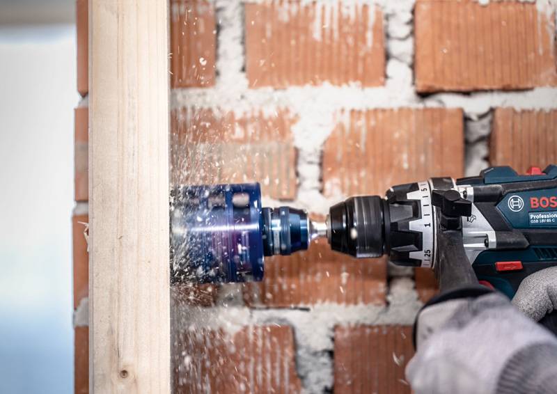 Ein Elektroschrauber mit Kreissägen-Aufsatz schneidet in Holz ein, erzeugt Sägespäne, vor einem Hintergrund einer freiliegenden Backsteinmauer.