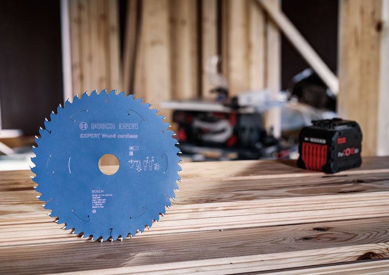 Ein Kreissägeblatt und ein Akkupack für Elektrowerkzeuge ruhen auf einer Holzplanke in einer Werkstattumgebung, mit unscharfen Werkzeugen im Hintergrund.
