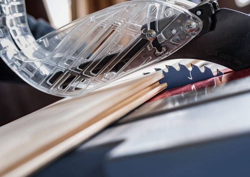 Nahaufnahme einer Kreissäge, die ein Holzbrett schneidet. Das Brett ist teilweise durchtrennt, und die durchsichtige Schutzvorrichtung über dem Sägeblatt ist deutlich sichtbar.