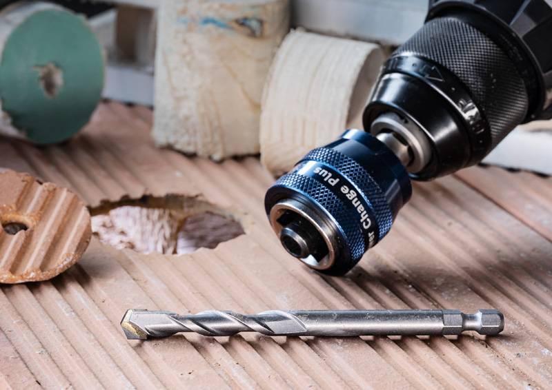 Ein Bohrmeißel und eine Bohrmaschine liegen auf einer Holzoberfläche mit Löchern. Die Szene deutet auf Holzbearbeitung oder Bauaktivität hin.