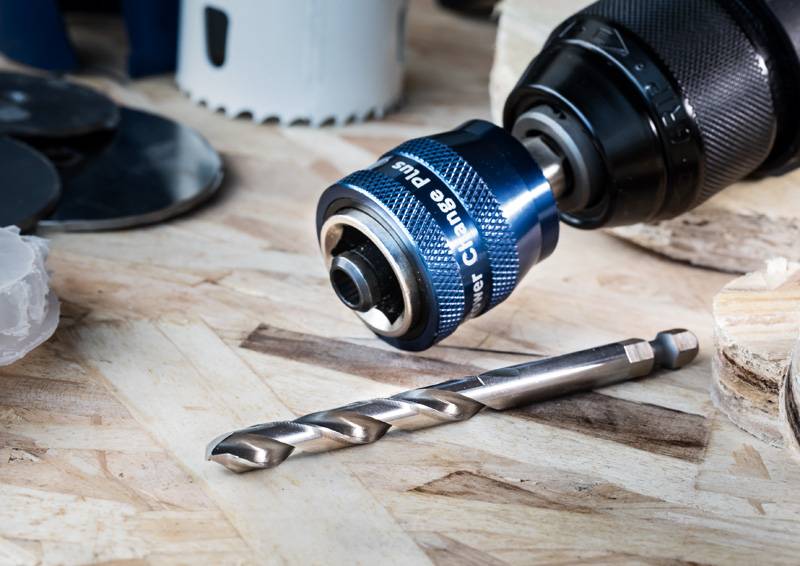Ein Bohrgerät mit einem blauen Schnellspannfutter und einem Metallbohraufsatz auf einer Holzoberfläche, umgeben von Bohrerzubehör und Werkzeugen.