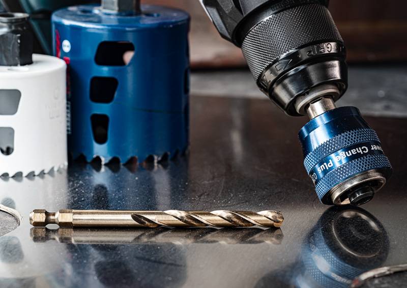 Ein Metallbohrer auf einer glänzenden Oberfläche platziert, mit einer blau-weißen Lochsäge im Hintergrund, die auf eine Werkstattumgebung hindeutet.