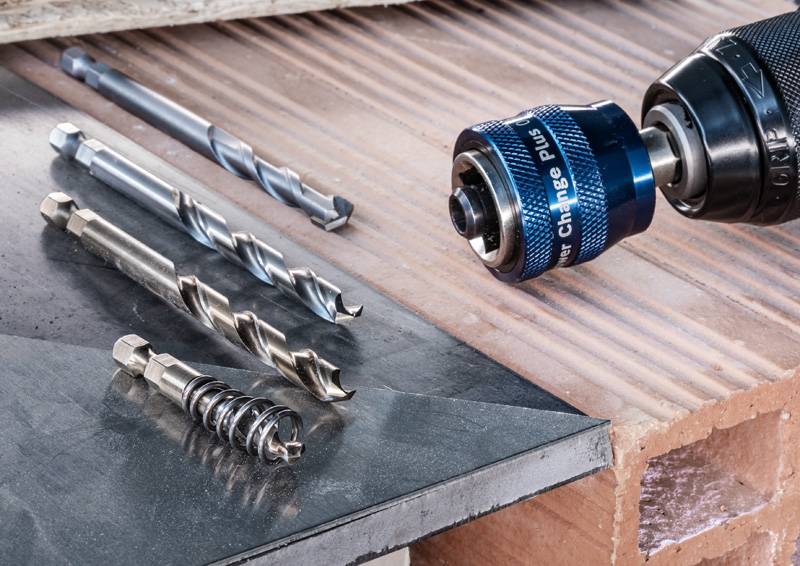 Ein Bohrer mit einem blauen Bohrfutter und verschiedene Bohrmeißel sind auf einer Holzoberfläche und einem schwarzen Metallblech angebracht, was auf eine Baustelle hindeutet.