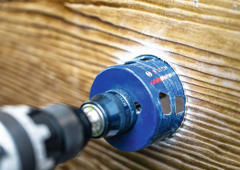 Eine blaue Lochsäge, die an einem Bohrer befestigt ist, schneidet durch ein Stück Holz und erzeugt dabei ein kreisrundes Loch.