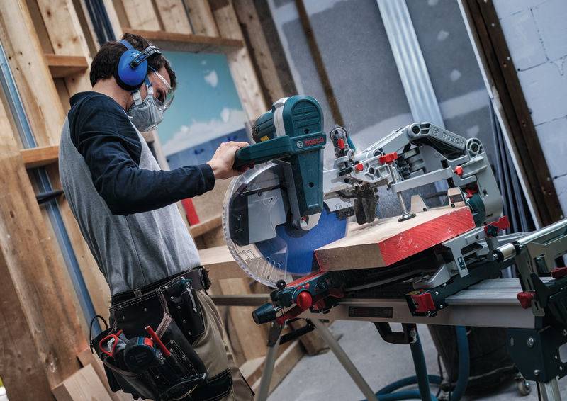 Person mit Schutzausrüstung bedient eine Kreissäge auf einem Holzbrett in einer Werkstattumgebung.