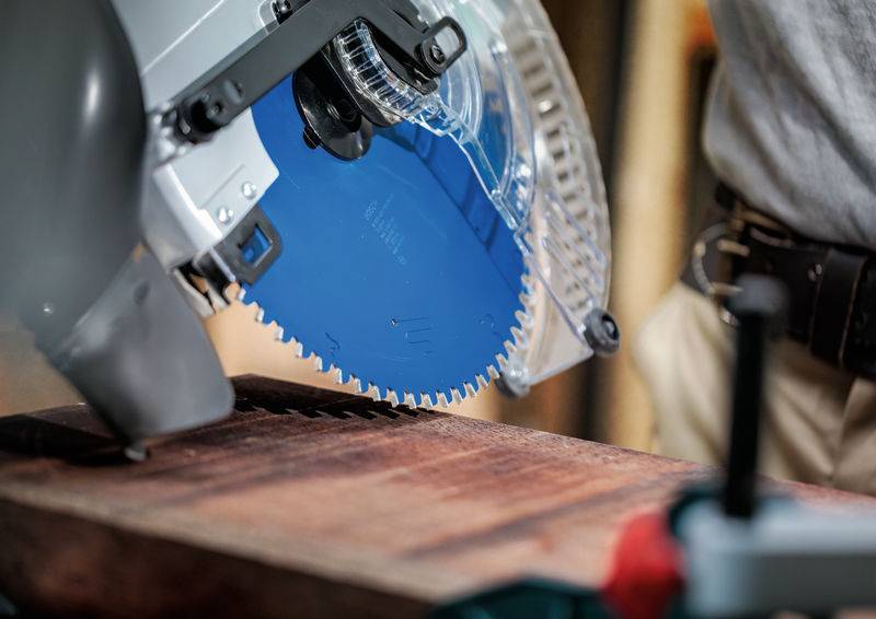 Eine Kreissäge mit einem blauen Blatt, das durch ein Holzbrett schneidet, bedient von einer Person, die ein graues Hemd und einen Werkzeuggürtel trägt.